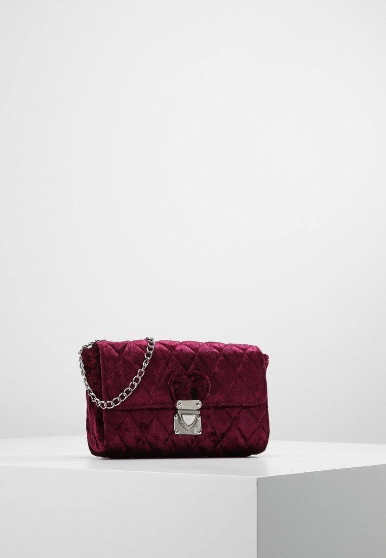 Bolso de mano de terciopelo acolchado en burdeos oscuro con una correa de cadena plateada y cierre. Presenta un acento en forma de corazón y una superficie texturizada.