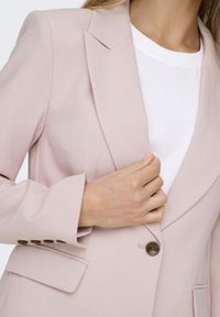Blazer rose pâle au design élégant, avec des revers crantés et un unique bouton marron. Comprend des poches latérales et des manches structurées portées sur un haut blanc.