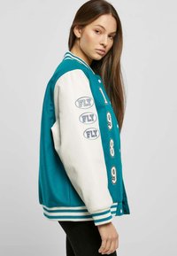 Veste varsity teal avec des manches en cuir blanc, patchs brodés 'FLY' et le numéro '1989' à l'avant. Comprend des poignets et un ourlet rayés.