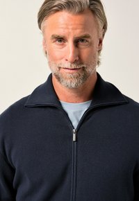 Giacca blu navy con zip, collo alto, tessuto liscio e cursore della zip argentato, indossata sopra una camicia azzurra.