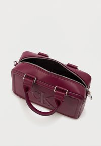 Burgundi värvi nahast kott ristküliku kujuga, jäädvustatud logo, kahekordne käepide ja hõbedane tõmblukk. Vooderdatud sisemus on nähtav.