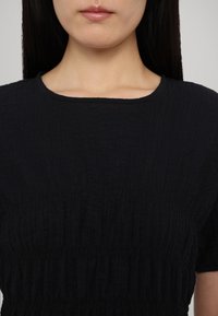 Haut noir texturé avec un col rond et des manches courtes. Présente un détail froncé le long du torse pour une coupe ajustée.