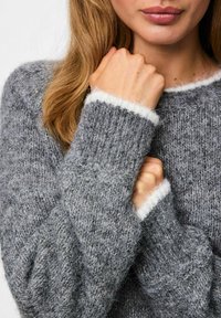 Femme vêtue d'un doux pull en tricot gris avec des finitions blanches sur les poignets, tenant ses poings près de sa poitrine, montrant le tissu texturé.