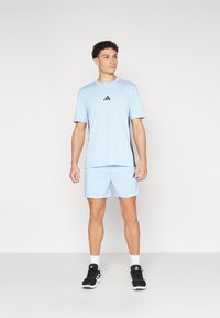 Šviesiai mėlynas sportinis marškinėlis ir šortai su juodu "adidas" logotipu, derinami su juodais bėgimo batais. Audinys atrodo lengvas ir pralaidus orui.