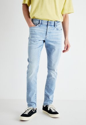 Person trägt hellblaue Slim-Fit-Jeans, gelbes T-Shirt und schwarze Sneakers mit weißen Schnürsenkeln, steht vor einem schlichten hellen Hintergrund.
