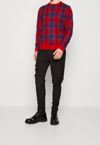 Rot-blau gemusterter Pullover mit Rundhalsausschnitt, kombiniert mit schwarzen Cargo-Hosen mit Seitentaschen und schwarzen Schnürstiefeln.