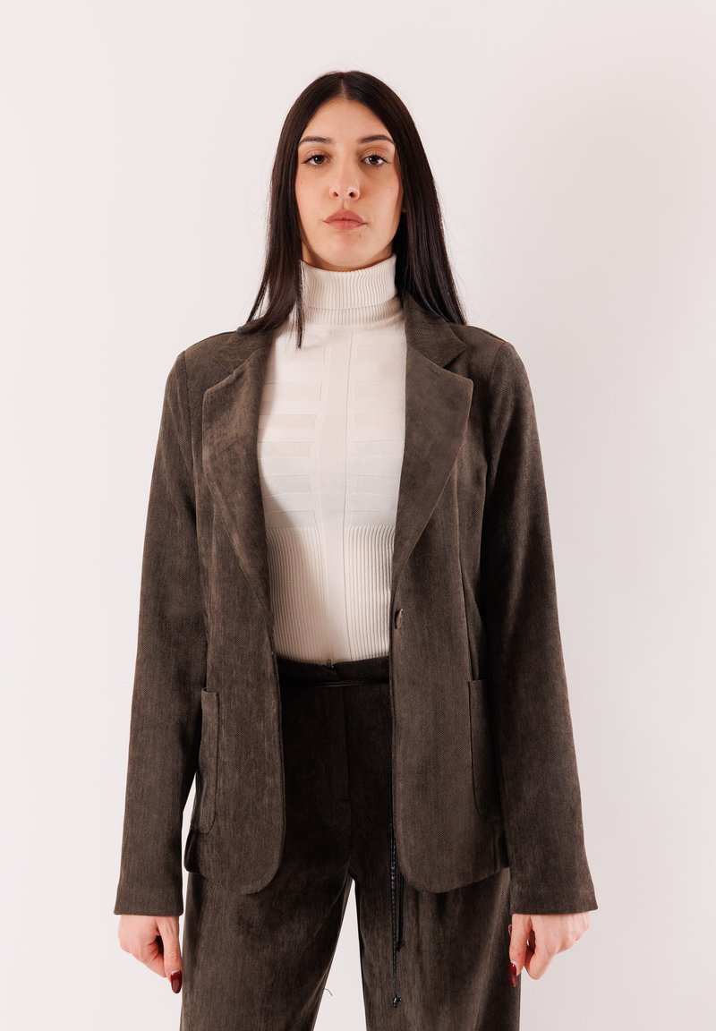 Blazer in velluto a coste verde oliva con bavero a lancia, tasche frontali e vestibilità morbida, abbinato a un dolcevita a coste bianco.