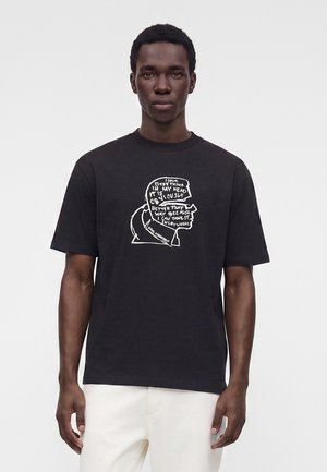 Mand iført en sort t-shirt med en hvid grafik af et hoved fyldt med håndskrevet tekst, kombineret med lyse bukser.