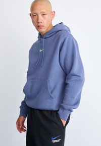 Blå hettegenser med kengurulomme, snor i hetten og en liten grønn Nike-logo på brystet; kombinert med svart joggebukse med blå tekst.