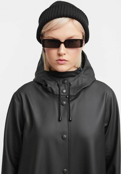 Zwarte waterdichte jas met capuchon, voorzien van drukknoopsluitingen en koordjes. Draag deze met een zwarte gebreide beanie en donkere zonnebril.