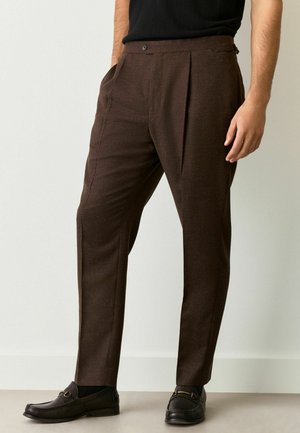Pantalones de vestir marrones con un tejido texturizado, que presentan bolsillos laterales, un cierre frontal con botón y un corte entallado. Llevados con mocasines negros.