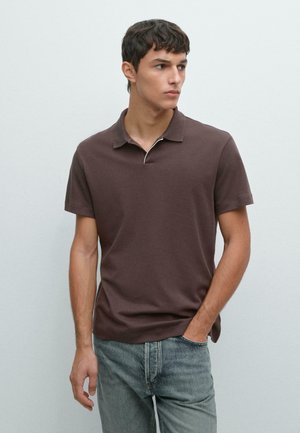 Jeune homme portant un polo marron à manches courtes et un jean bleu clair, debout avec une main dans la poche, regardant sur le côté.