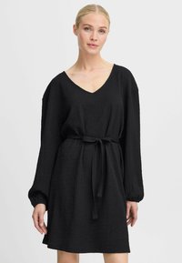 Robe noire en tissu texturé, présentant un décolleté en V, des manches longues et une ceinture nouée à la taille pour un ajustement réglable.