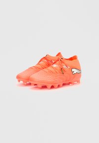 Zapatillas de fútbol de color naranja brillante con superficies texturizadas, diseño con cordones y un logo blanco en el costado. Presentan tacos moldeados para mayor tracción.