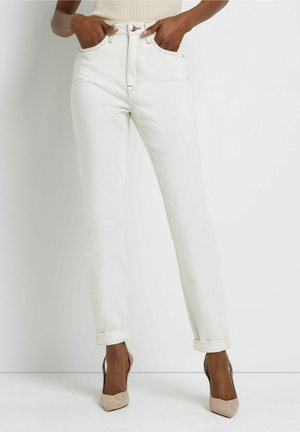 Naismalli, jolla on päällään valkoiset korkeavyötäröiset farkut, joissa on käännetyt lahkeet, ja beiget teräväkärkiset slingback-korkokengät, kädet taskuissa valkoista taustaa vasten.