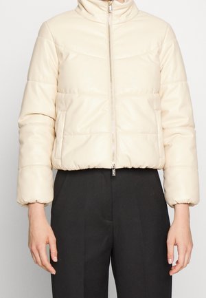 Chaqueta acolchada corta de color crema con cremallera frontal y bolsillos laterales, usada sobre pantalones negros a la cintura alta y de corte entallado.