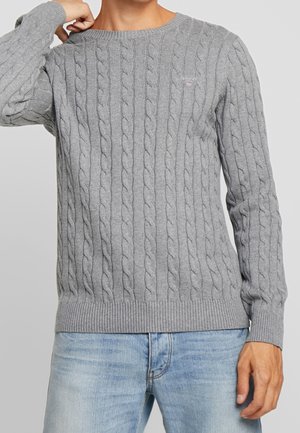Man wearing a light gray cable-knit sweater with ribbed cuffs and hem, paired with light blue jeans against a plain background.
