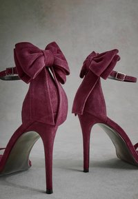 Sandales à talons hauts en daim bordeaux avec de grands nœuds à l'arrière, des brides fines autour de la cheville et des bouts pointus, dotées d'une doublure intérieure lisse.