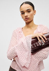 Ροζ πουά μπλούζα με λαιμόκοψη με βολάν και μανίκια με φούντα. Το μοντέλο κρατάει μια σκούρα κόκκινη τσάντα χειρός με ασημένιο μεταλλικό εξάρτημα και οπές.