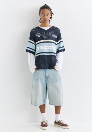Junge Person mit geflochtenem Haar, die einen dunkelblauen Netzpullover über weißen langärmeligen Kleidungsstücken, hellblaue Jeansshorts, weiße Socken und braune Turnschuhe trägt.