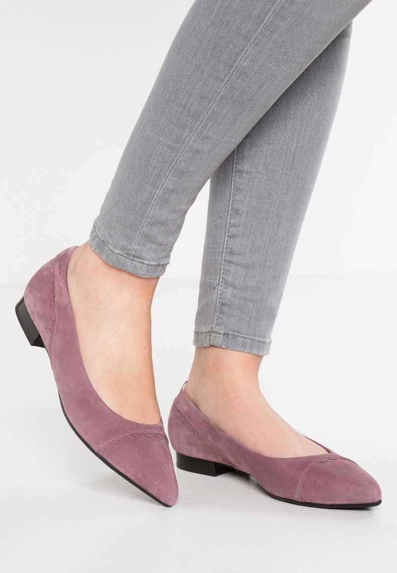 Ballerines pointues en daim mauve usé à petit talon, associées à un jean gris clair ajusté, sur un fond blanc uni.