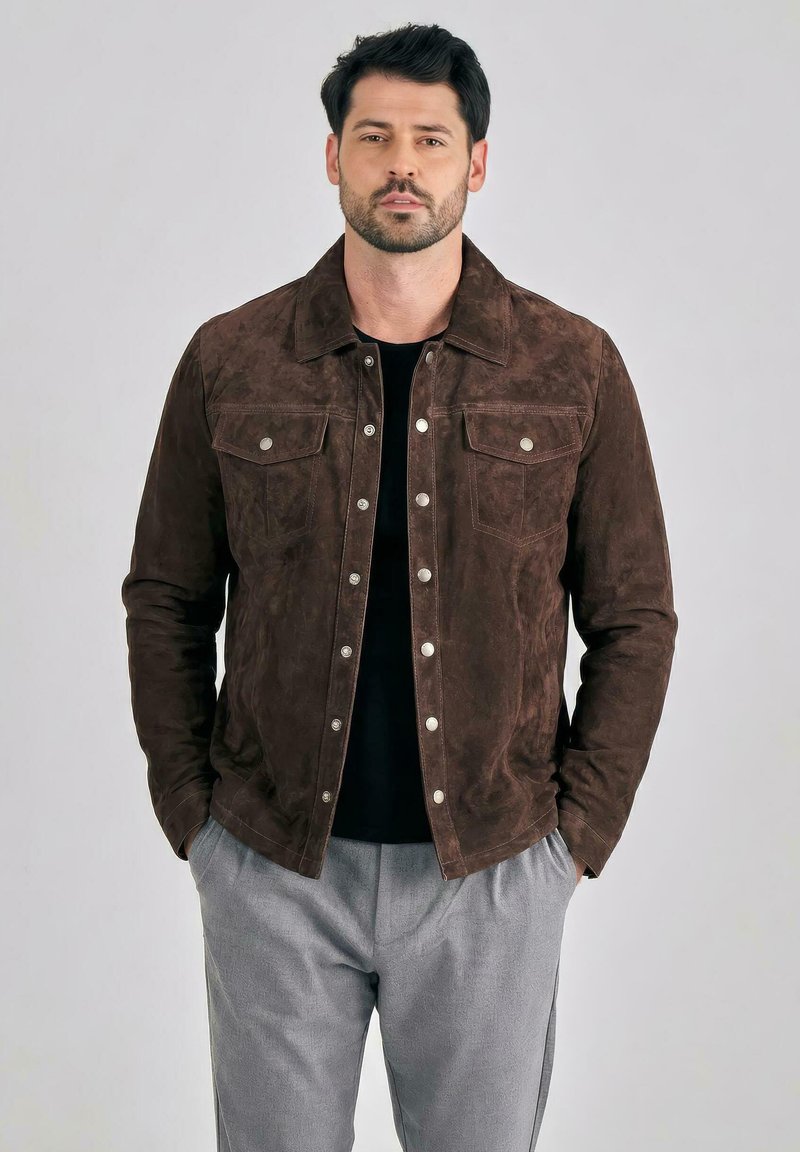 Mann trägt eine dunkelbraune Wildlederjacke mit silbernen Knöpfen über einem schwarzen Hemd und hellgraue Hose, steht mit den Händen in den Taschen.