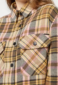 Gros plan d'une chemise à carreaux rose, marron, jaune et grise, avec des poches poitrine à boutons et des boutons noirs.