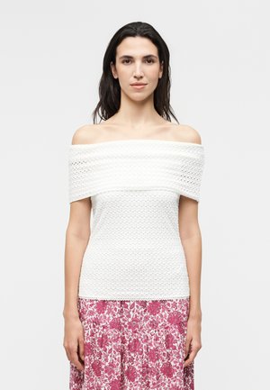 Γυναίκα με σκούρα μαλλιά φοράει ένα λευκό υφασμάτινο τοπ με ακάλυπτους ώμους και μια κόκκινη φούστα με λουλουδένιο σχέδιο, στέκεται μπροστά σε μια απλή φόντο.