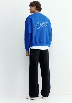 Jeune homme portant un sweat-shirt bleu avec un texte graphique blanc dans le dos, un jean noir à jambes larges et des chaussures blanches sur fond blanc uni.
