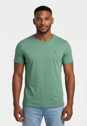 Mann trägt ein eng anliegendes grünes T-Shirt mit kurzen Ärmeln und hellblaue Jeans, steht vor einem schlichten weißen Hintergrund.