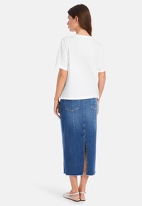 Weißes Baumwoll-T-Shirt mit kurzen Ärmeln, kombiniert mit einem blauen Jeansrock mit einem Schlitz hinten und fransigem Saum. Sandalen mit Riemchen an den Füßen.