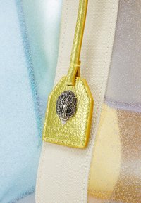Étiquette en cuir jaune avec un emblème décoratif en métal attaché à une sangle tissée beige sur un fond doux bleu et jaune.