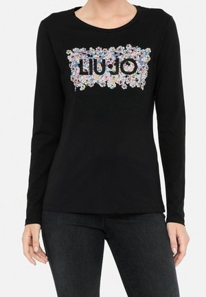 Vrouw die een zwart shirt met lange mouwen draagt met een bloemenapplicatie en de tekst "LIU JO" in het midden op de borst.