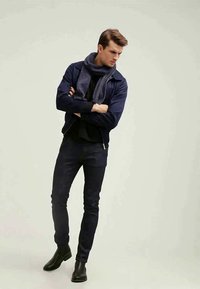 Casaco de piloto azul-marinho, gola alta preta, jeans escuros, cachecol cinza. Botas de cano curto pretas. Materiais suaves, corte justo, design minimalista.