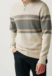 Maglione beige con zip al collo, strisce orizzontali nere e marroni, tessuto testurizzato, polsini e orlo a coste. Abbinato a pantaloni scuri.