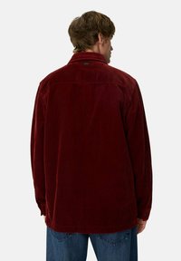 Mand, der står med ryggen til, iført en løs, langærmet, mørkerød fløjlsskjorte og blå denimjeans foran en ensfarvet baggrund.