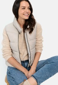 Helle beige Pufferweste mit hohem Kragen, Reißverschluss vorne und gestepptem Material. Kombiniert mit einem cremefarbenen Strickpullover und blauen Jeans.