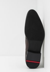 Scarpa in pelle marrone con una suola in gomma nera testurizzata, caratterizzata da un accento rosso sul tallone e un design elegante e lucido.