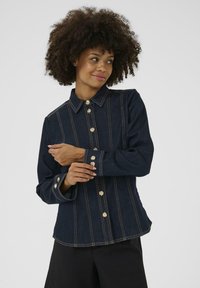 Žena s kudrnatými vlasy, oblékající tmavě modrou džínovou bundu se zlatými knoflíky, která si upravuje manžetu, na jednobarevném světlem pozadí.