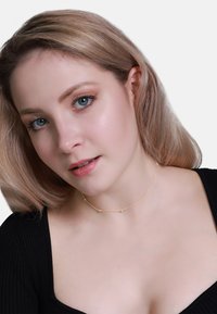 Gouden choker ketting met kleine ronde accenten, fijn kettingontwerp, gedragen met een zwarte geribbelde top. Zachte focus op huidtint en gelaatstrekken.