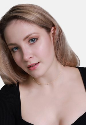 Collana choker d'oro con piccoli accenti tondeggianti, design di catena fine, indossata con una maglia nera a coste. Sfocatura lieve sul tono della pelle e sui tratti del viso.
