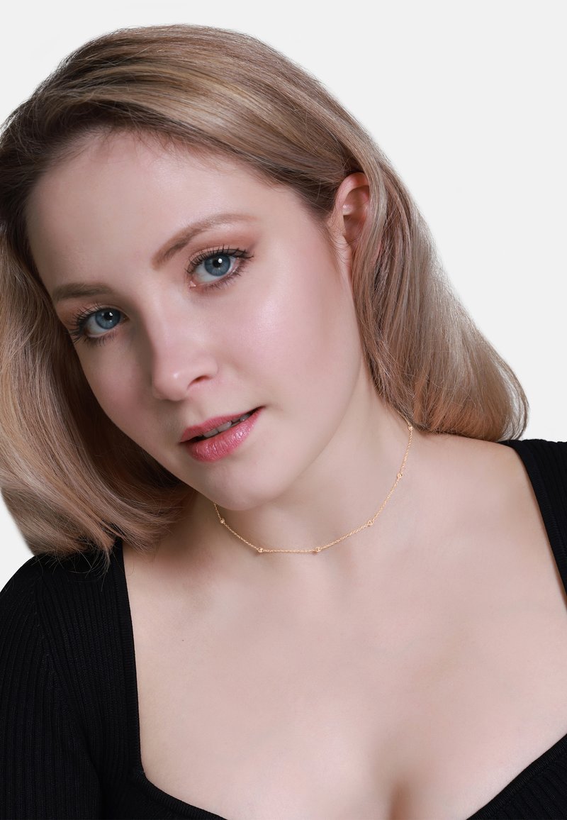 Gouden choker ketting met kleine ronde accenten, fijn kettingontwerp, gedragen met een zwarte geribbelde top. Zachte focus op huidtint en gelaatstrekken.