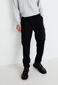 Svart cargo bukser med elastisk liv, sidelommer og avslappet passform, kombinert med blanke svarte sko. Stoffet ser jevnt og slitesterkt ut.