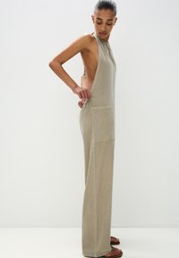 Hellbeige ärmelloser Jumpsuit mit hohem Kragen, großen seitlichen Ausschnitten, zwei Fronttaschen und einem lockeren, entspannten Schnitt. Weicher Stoff.