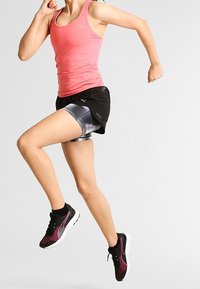 Camiseta sin mangas rosa, pantalones cortos para correr en negro y plateado, zapatillas deportivas negras y rosas. Sujetando una pose de carrera contra un fondo liso.