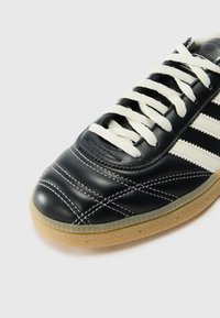 Black leather sneaker with white laces and three white stripes. Features detailed stitching and a textured gum sole.