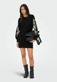 Zwart gebreid jurk met lange mouwen, versierd met witte bloemenborduursels, gecombineerd met een zwarte gestructureerde handtas en zwarte laarzen tot de knie.