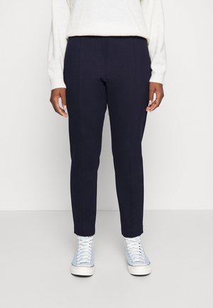 Person trägt marineblaue, maßgeschneiderte Hose, weißen Pullover und hellblaue High-Top-Sneaker, steht vor einem schlichten weißen Hintergrund.