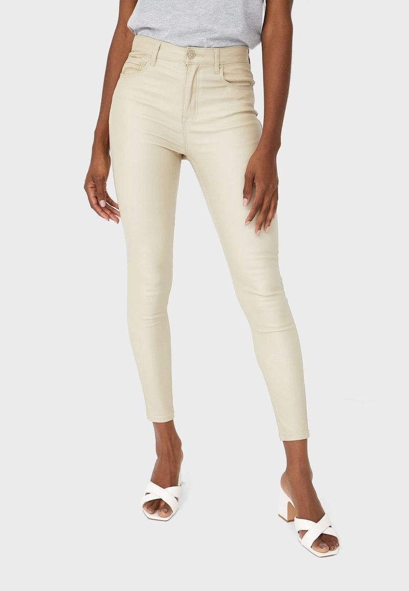 Jean skinny taille haute beige avec une texture lisse et un minimum de détails, comprenant des poches avant et une fermeture éclair. Associé à des sandales blanches à talons blocs.