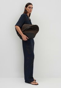 Sac en tissu tissé brun avec une base noire, au design courbé, tenu par une personne vêtue d'une tenue marine foncé ornée de lignes abstraites, portant des sandales plates.